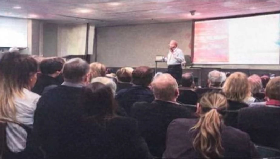 Professional presentation to a crowded auditorium with a speaker at a podium and projection screen visible