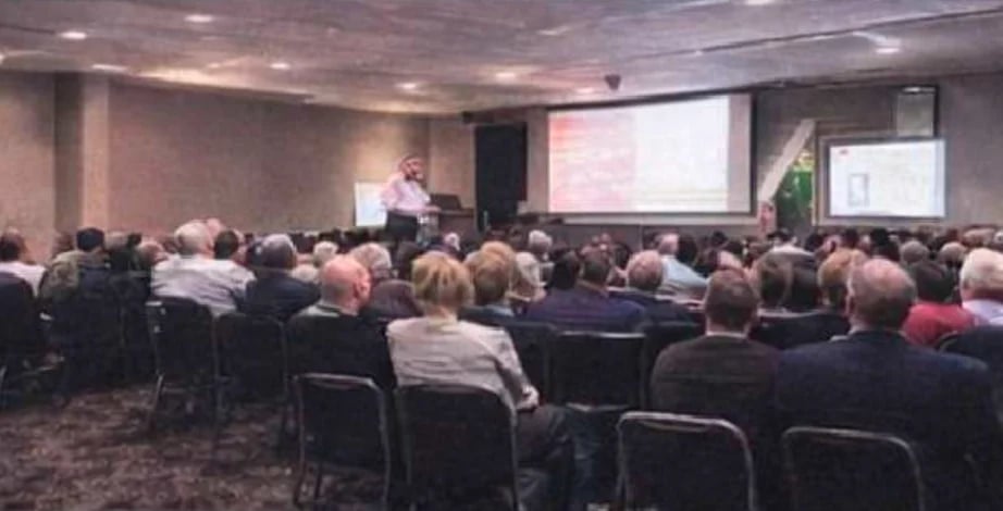 Large audience seated in rows facing a presenter at a projector screen during an indoor conference or seminar